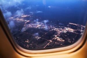 The image shown is that of an aerial view of a city landscape at night through the window of an airplane. Lights can be seen against a dark blue, night sky. Several clouds are seen on the left hand side of the image. 